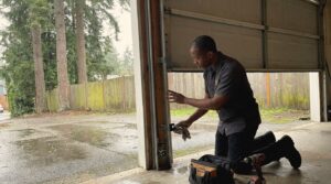 Perfection Garage Door technician wiping a fogged photo-eye safety sensor at the base of a garage door track in Renton, WA on a rainy day