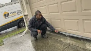 Perfection Garage Door technician crouched at the bottom of a garage door in the rain at a Renton, WA home, checking where water runs under the door, company van parked behind
